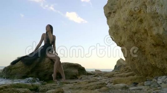 一个忧郁的黑发女人穿着一件通风的黑色连衣裙，坐在一块破碎的岩石上，海浪在盐滩上拍打着。视频