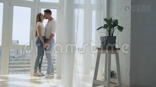 年轻的幸福夫妇在他们的新公寓的窗户附近拥抱和亲吻视频