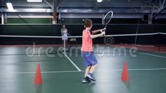 穿着白色运动服的女人正在室内球场和她的儿子打网球。 女教练在服役，男孩在视频