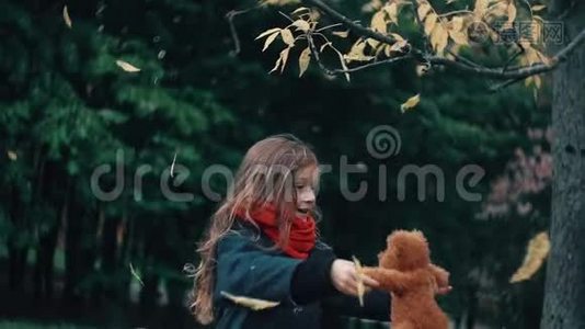 有趣的，欢快的，可爱的小女孩带着她的玩具旋转，泰迪熊黄色的叶子从树上慢慢地落在它们身上。视频