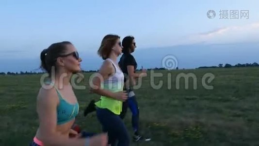 美丽的，运动的，年轻的女人戴着太阳镜，穿着紧身衣，沿着绿色的草地，田野同步奔跑。 在夏天视频