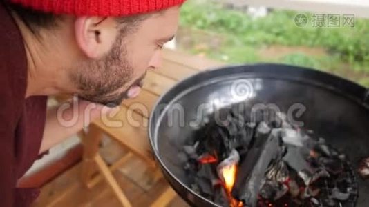 年轻的大胡子在烧烤炉里吹着煤火. 黑色木制木炭燃烧在Bbq。 高清高清视频