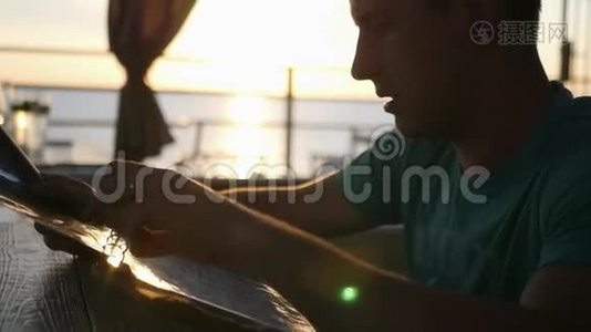 一个年轻人坐在咖啡馆里，在地平线上美丽的日落背景下，从菜单中挑选食物视频