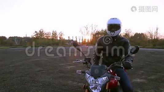 一个穿着黑色皮夹克和白色头盔的年轻人坐在摩托车上，在秋日日落前开灯视频