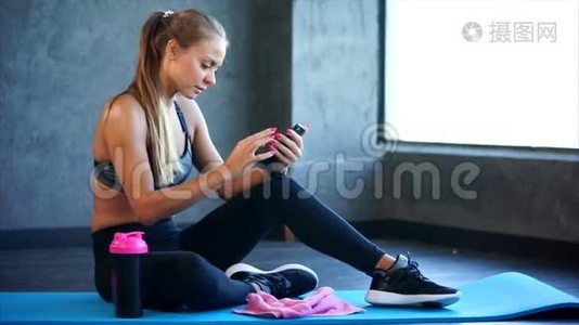 健身房里有智能手机的女人。 她坐在垫子上用智能手机视频