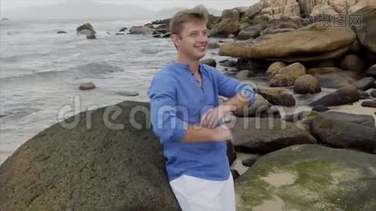 一个穿着蓝色衬衫的微笑的金发男人站在大石头附近的海滩上，理直了头发视频