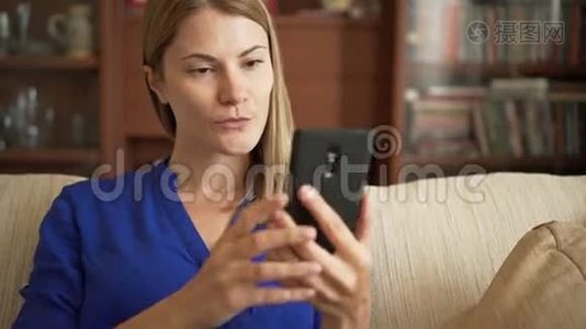 漂亮的年轻女士穿着蓝色衬衫，坐在客厅的沙发上，用智能手机自拍视频