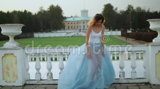 漂亮的瘦女孩穿着白色和蓝色的连衣裙站在白石栏杆附近，在照片拍摄时微笑和摆姿势。视频