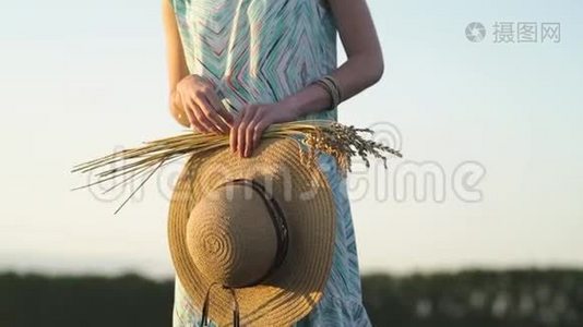 戴太阳镜的女孩站在日落时的田野里，拿着金色的麦穗和草帽。 乡村户外场景视频