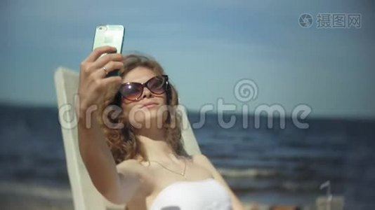 一个穿着白色比基尼的年轻女孩躺在海边沙滩上的休息室里晒太阳，并使用智能手机视频
