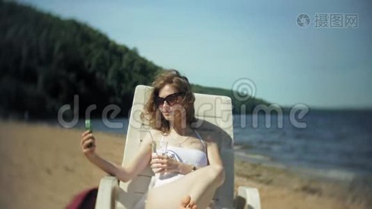一个穿着白色比基尼的年轻女孩躺在海边沙滩上的休息室里晒太阳，并使用智能手机视频
