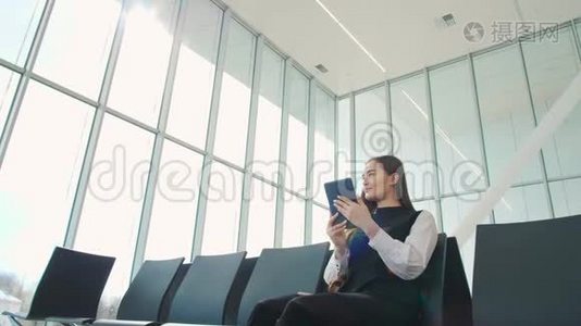 年轻的女乘客在机场，一边用平板电脑一边等着她的航班，微笑着，阳光明媚的一天视频