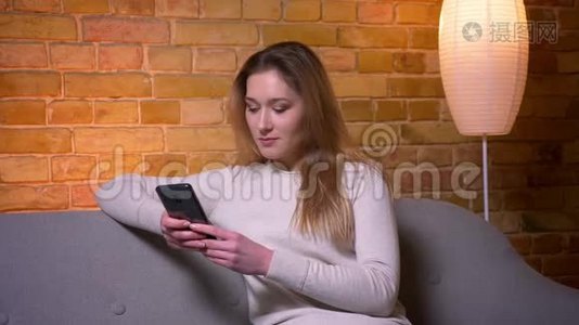 年轻漂亮的白种人深色头发的女性用电话坐在舒适的室内沙发上的特写镜头视频