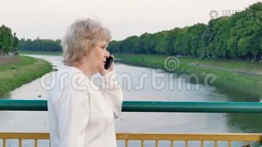 一位年长的女士穿过河上的桥，在智能手机上交谈视频