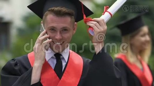 微笑的男毕业生拿着大学文凭，在电话里交谈视频
