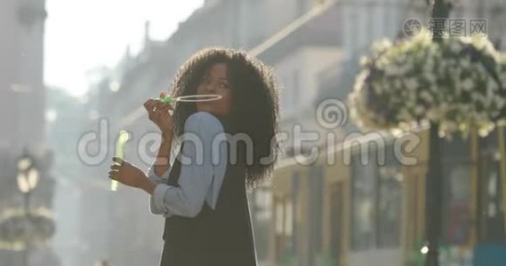 迷人的非洲裔美国女孩在肩膀上吹肥皂泡的侧视。 街道的位置。视频