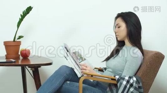 漂亮的女孩坐在舒适的椅子上，在家看一本光滑的杂志视频