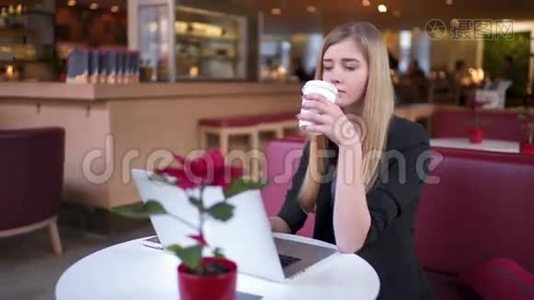 漂亮的年轻女孩，穿着黑色夹克的金发坐在咖啡馆A©里。 她喝咖啡和工作视频