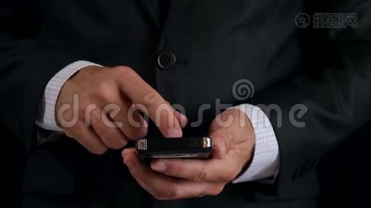 一个穿黑色西装的商人在网上工作。 一个男人的手拿着一部黑色背景的时尚手机。视频