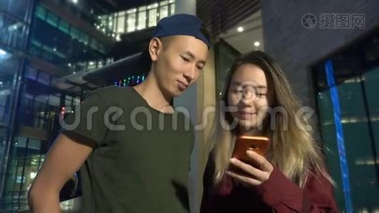 快乐的年轻亚洲夫妇的男朋友和女朋友。 晚上站在城市街道上使用智能手机。视频