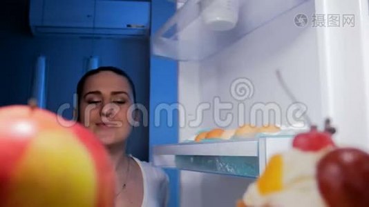 4K镜头，年轻微笑的女人在夜里看着冰箱里，咬着红苹果视频