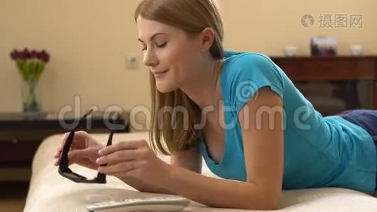 美丽迷人的年轻女子穿着蓝色t恤躺在沙发上，穿着3D眼镜看电视。视频