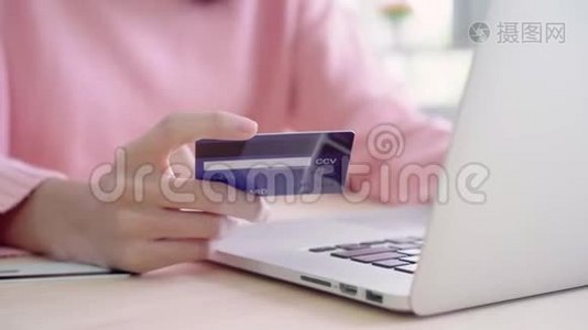 美丽的亚洲女人用电脑或笔记本电脑通过信用卡购买网上购物，而穿毛衣坐在桌子上。视频