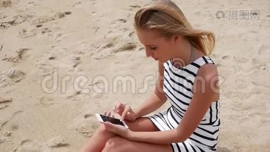 年轻漂亮的女士身材苗条，一头金发，穿着黑白连衣裙，坐在海滩上，使用智能手机视频