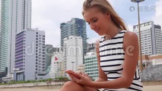 年轻漂亮的女士身材苗条，一头金发，穿着黑白连衣裙，坐在海滩上，使用智能手机视频