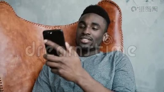 非洲年轻人坐在椅子上的肖像，使用智能手机。 一个英俊的男人微笑着在电话里说话。视频