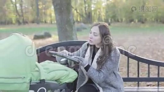 秋天。 一位年轻的母亲坐在公园的长凳上摇着婴儿车。视频