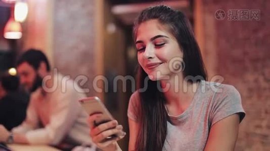 年轻迷人的女人，美丽的微笑是使用智能手机在晚上。 坐在酒吧或餐馆里的女孩视频