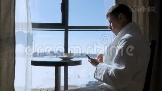 一个年轻有魅力的男人坐在靠窗的桌子旁喝茶，喝咖啡，在手机上写短信视频