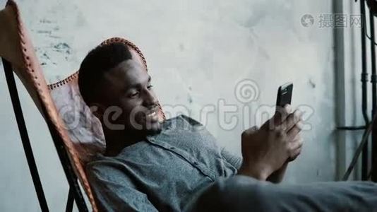 非洲男子坐在椅子上的肖像，使用智能手机。 英俊的男性微笑，看着手机里的照片。视频