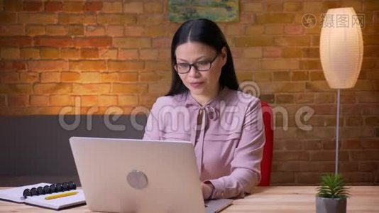 在办公室里，一位在笔记本电脑上工作、看着相机、微笑的成年亚洲女商人的特写镜头视频