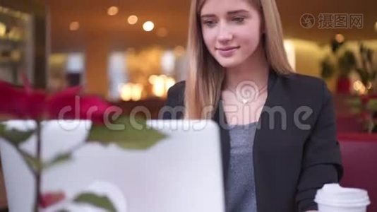 穿黑色夹克的金发女孩微笑着坐在咖啡馆里。 她正在喝咖啡，和她一起工作视频