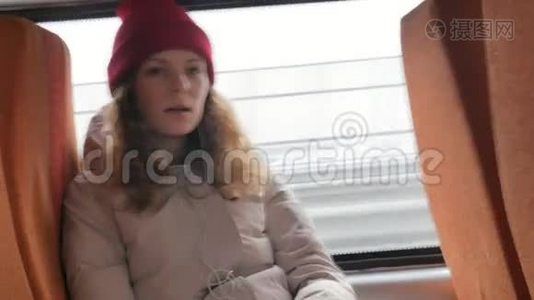 年轻开朗的女人戴着红帽子，在一个悲伤的日子乘公共汽车旅行。 她听音乐视频