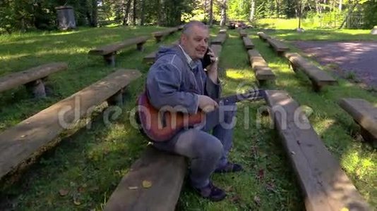 一个拿着吉他的人在电话里说话，然后在长凳上的公园里开始弹吉他视频