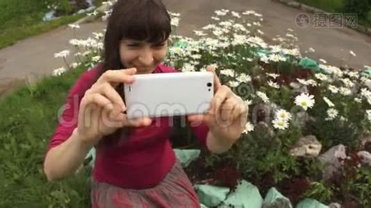 年轻女子坐在盛开的雏菊旁，用白色手机自拍。视频