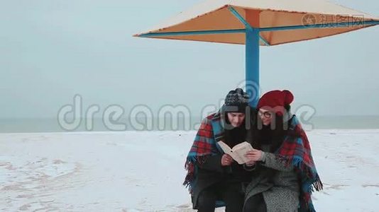 一个女孩和一个男人坐在海滩的天篷下，盖着毯子，看书。视频