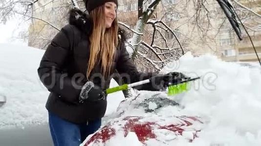 4k视频美丽微笑的年轻女子用刷子从雪地上清洁汽车挡风玻璃视频
