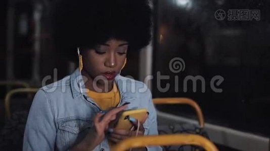 漂亮的非裔美国年轻女性在公共交通工具上使用智能手机。 晚上。 城市灯光背景。视频