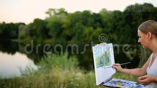 年轻艺术家在公园里用画笔和色彩坐在浪漫的湖泊和城市建筑旁。视频