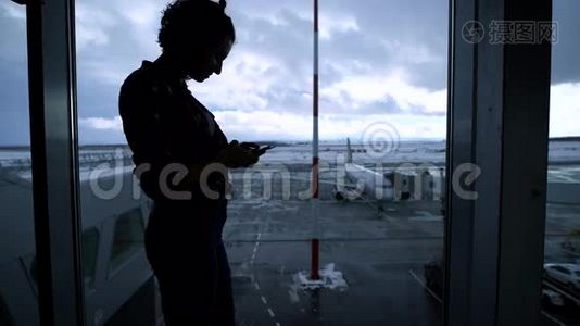 站在机场窗口附近使用智能手机的年轻健康女孩的剪影。 年轻的商业女性在等待视频