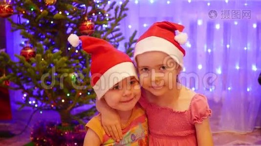 两个可爱的女孩微笑着，戴着圣诞帽对着镜头挥手。 背景，圣诞杉木的灯光和花环视频
