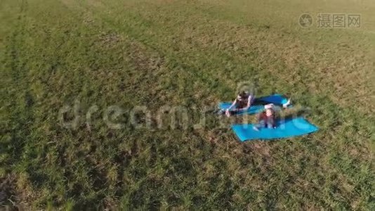 在温暖的夏日里，妈妈和她的小女儿在田野上的垫子上伸展。 空中全景。视频