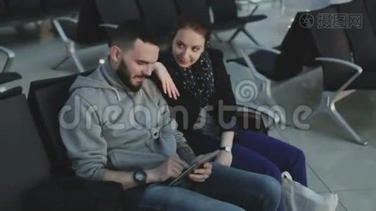 那家伙和穿着休闲服装的女孩看着坐在机场候机大厅的黑色椅子上的平板电脑。视频