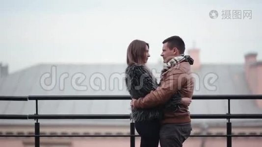 可爱的夫妇在寒冷的天气里站在屋顶上亲吻和大笑。 他们互相拥抱，穿着毛皮外套的女孩，侧视视频