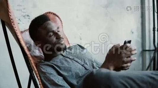 非洲年轻人坐在椅子上的肖像。 男正认真思考，手持智能手机.. 男人打字留言，微笑。视频