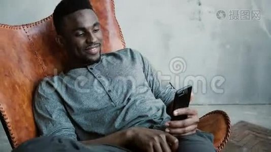 非洲男子坐在椅子上的肖像，使用智能手机。 人读留言，笑.. 打字按摩，微笑着视频